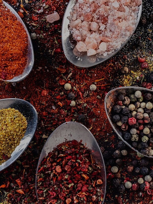 Spoons of paprika, pink salt, mixed peppercorns, and red pepper flakes on a spice-covered surface